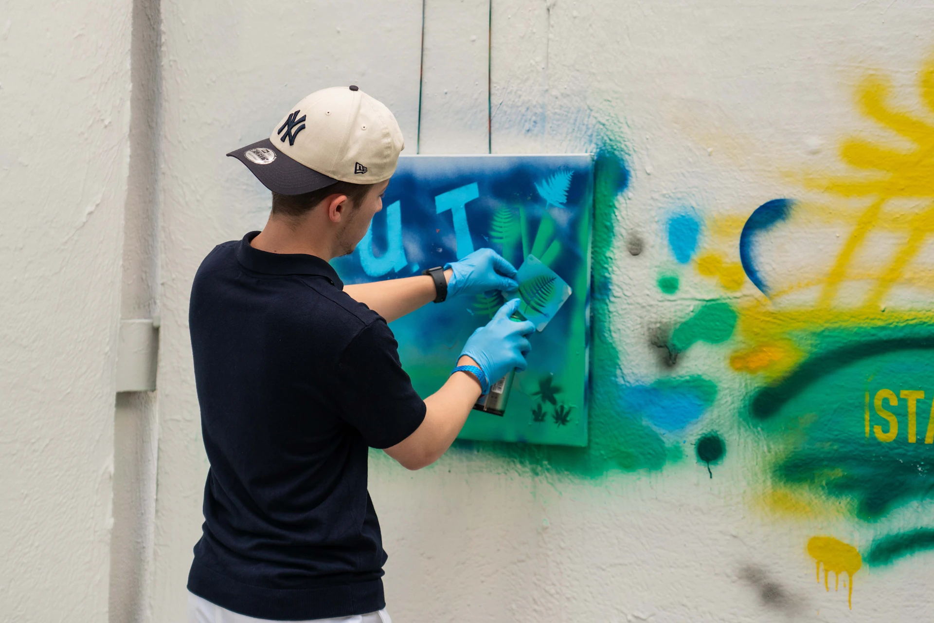 Person in a baseball cap and gloves spray-painting a stencil on a wall with colorful graffiti.