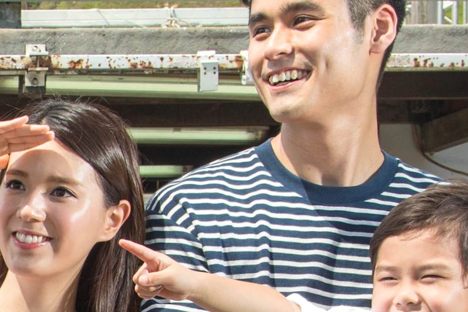 A smiling family of three poses on a sunny pier with mountains in the background; they stand behind a lifebuoy.