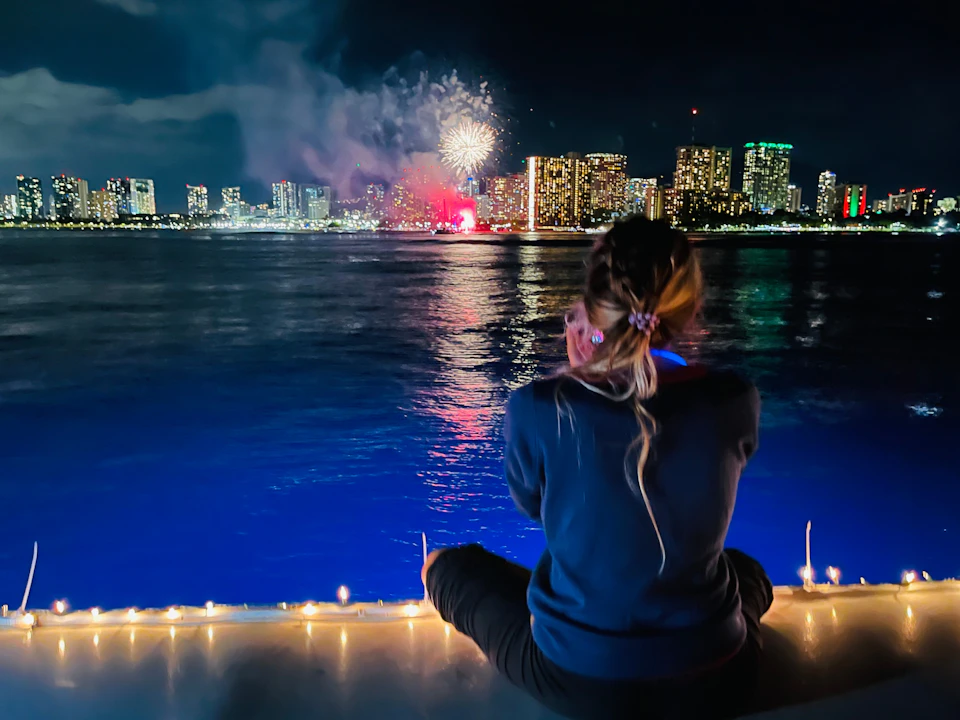 A person sitting on a ledge overlooking a city skyline at night, with fireworks illuminating the sky over the water.