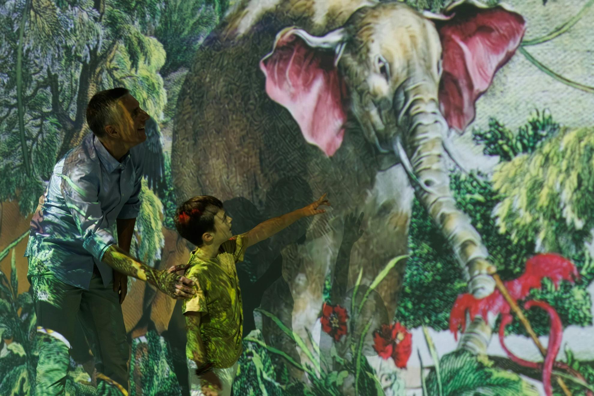 A child points at a projector image of an elephant with pink ears as an adult stands beside him, both illuminated by the projection.
