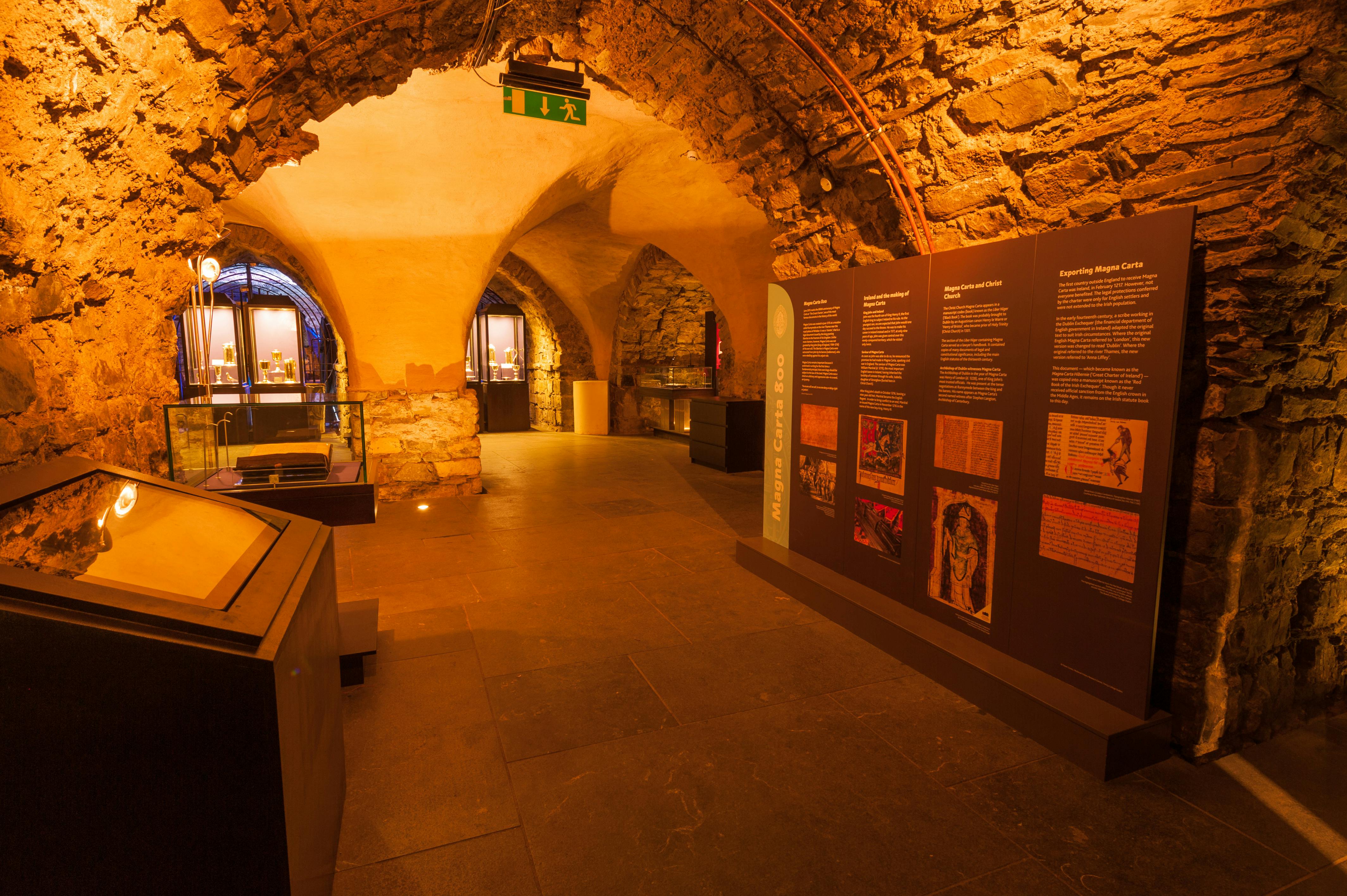 Una sala espositiva del museo in pietra con pannelli informativi, un arco, vetrine e un cartello di uscita verde in alto.
