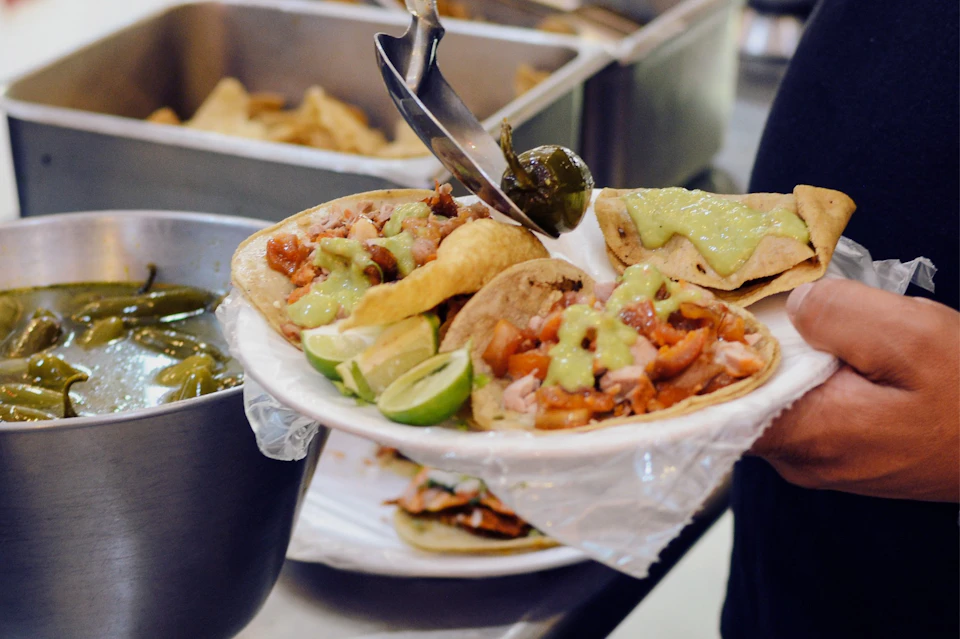 A plate of freshly prepared tacos topped with avocado salsa and lime wedges is served with a pickled jalapeño.