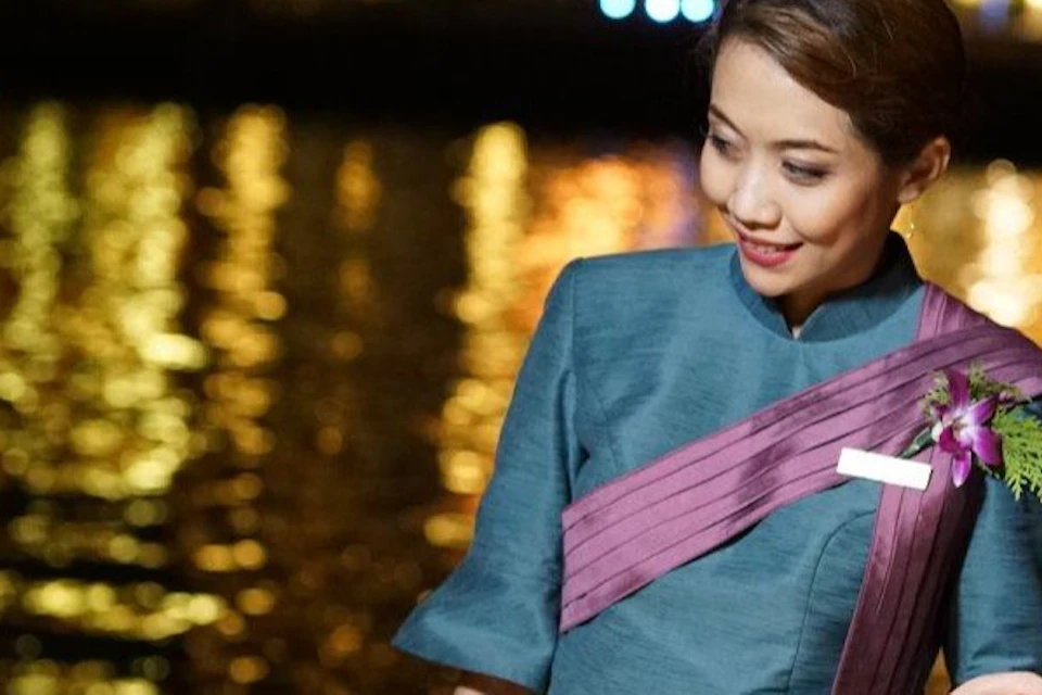 A man dines at a riverfront restaurant, conversing with two staff members dressed in traditional attire, under evening lights.