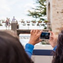 Person holding a smartphone, taking a photo of a bridge with people and flower pots on it, while another person watches.