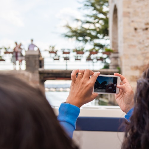 Person holding a smartphone, taking a photo of a bridge with people and flower pots on it, while another person watches.