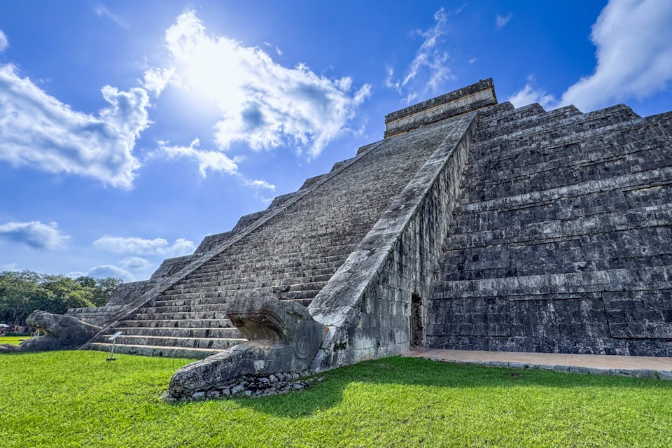 Stone pyramid with a stairway flanked by serpent sculptures, set against a blue sky with clouds. Green grass surrounds the base.