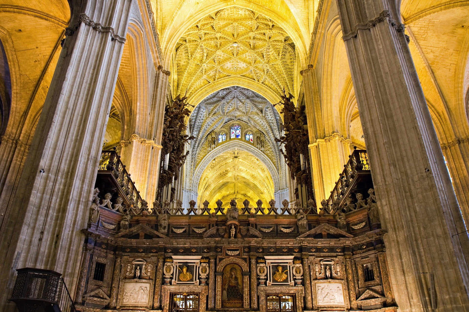 Interior of a cathedral with intricate stone carvings, tall columns, and a vaulted ceiling illuminated with golden light.