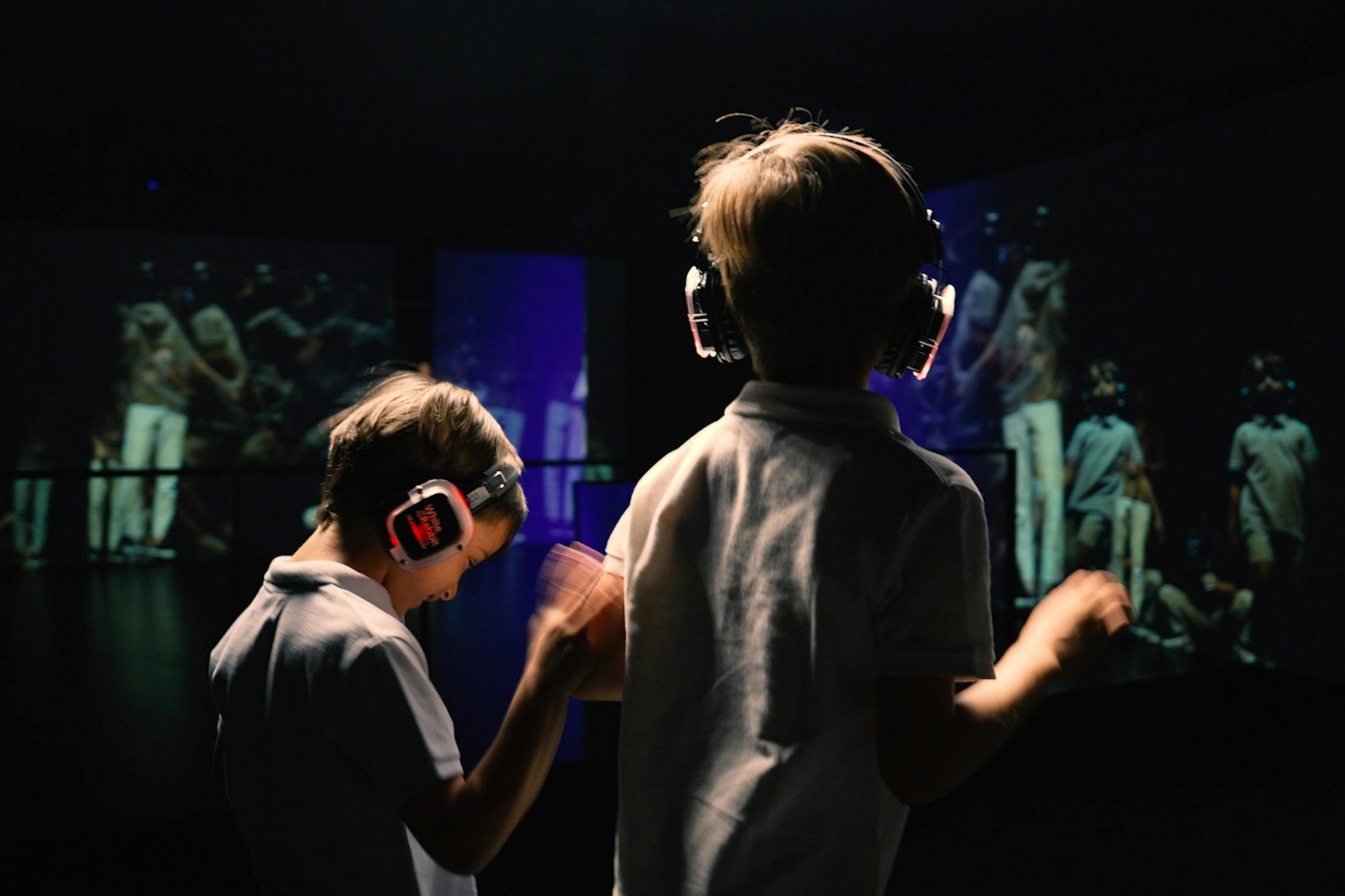 Deux enfants portant des écouteurs dansent dans une pièce sombre avec des écrans colorés affichant des personnes en arrière-plan.