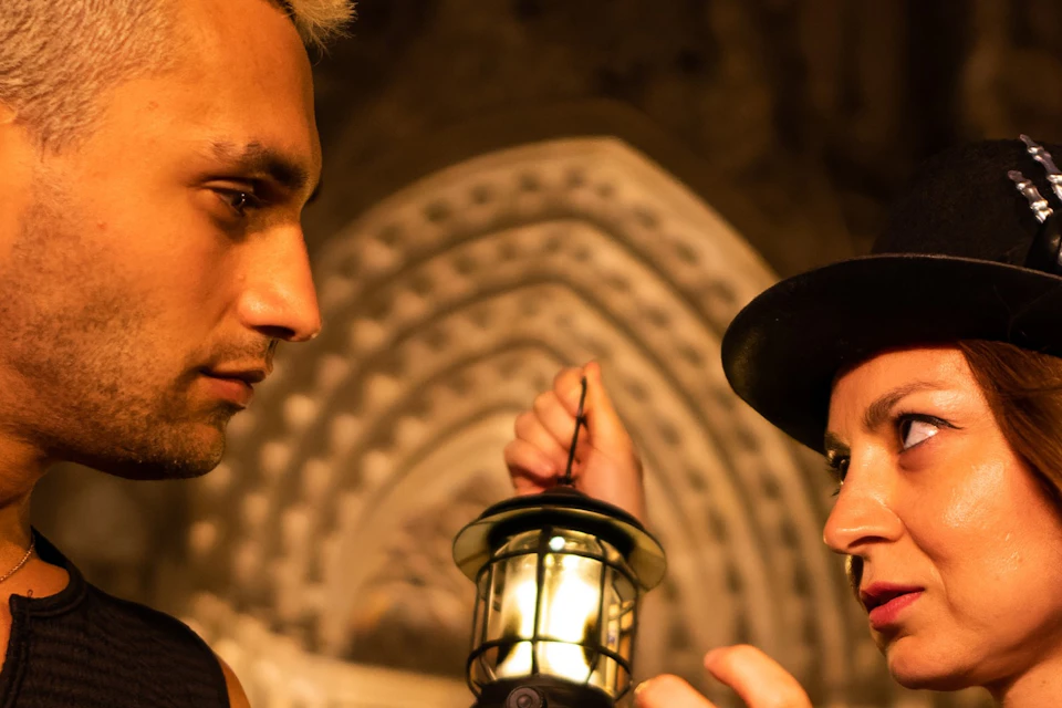 Two people face each other; one holds a lantern. They stand in front of an ornate, illuminated building at night.