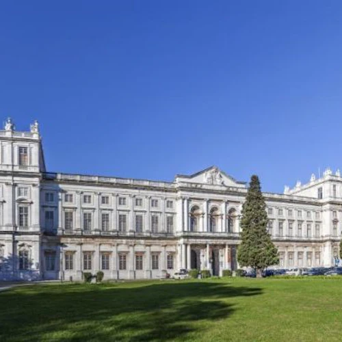 Stor, vit neoklassisk byggnad med flera fönster och en storslagen entré, belägen mitt i en välskött gräsmatta under en klarblå himmel.