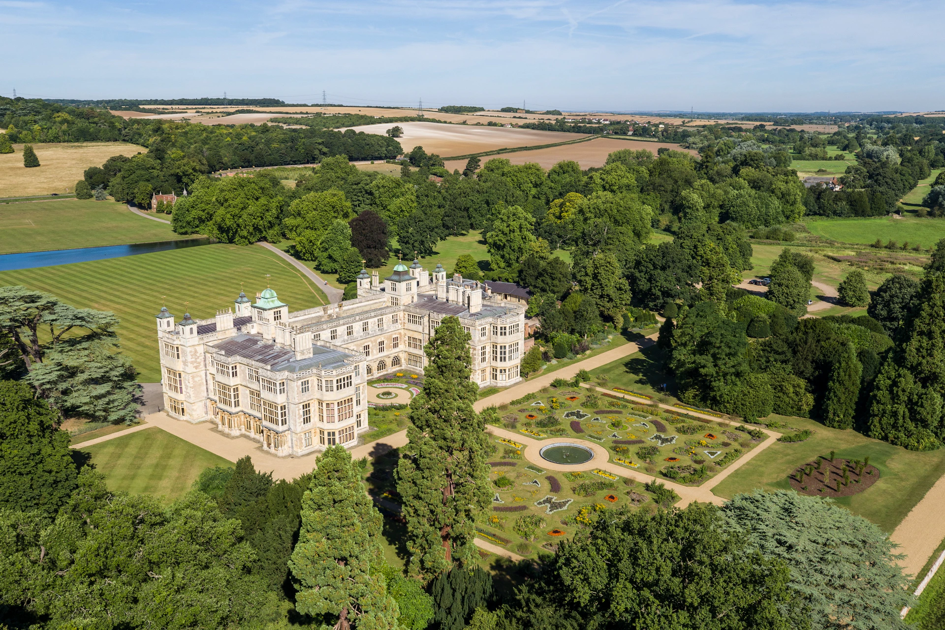 Aerial view of a large historic mansion surrounded by formal gardens and lush greenery, with fields in the background.