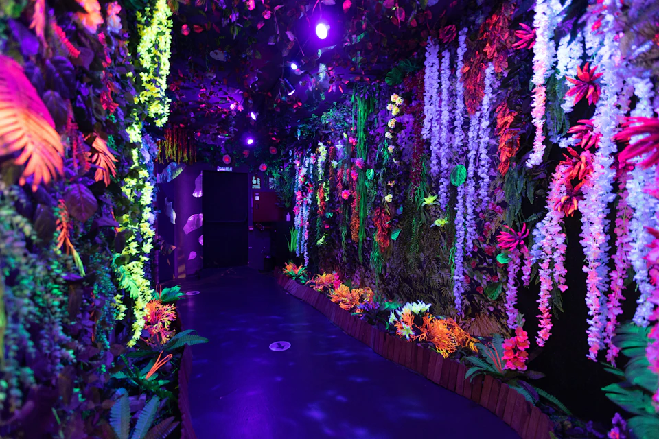 A dimly lit hallway adorned with colorful, glowing artificial flowers and foliage, illuminated by purple and blue lights.