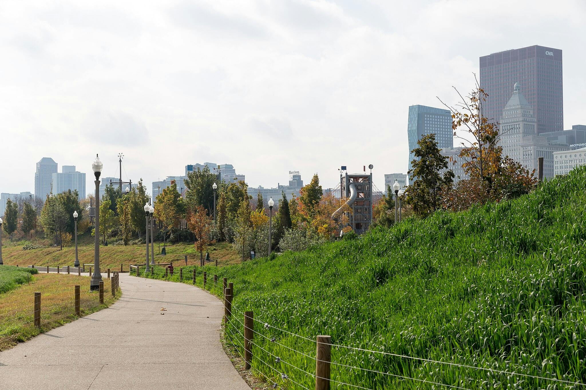 Sentier sinueux traversant un parc avec des lampadaires, des arbres et des collines herbeuses, menant à la ligne d'horizon d'une ville avec de grands bâtiments.