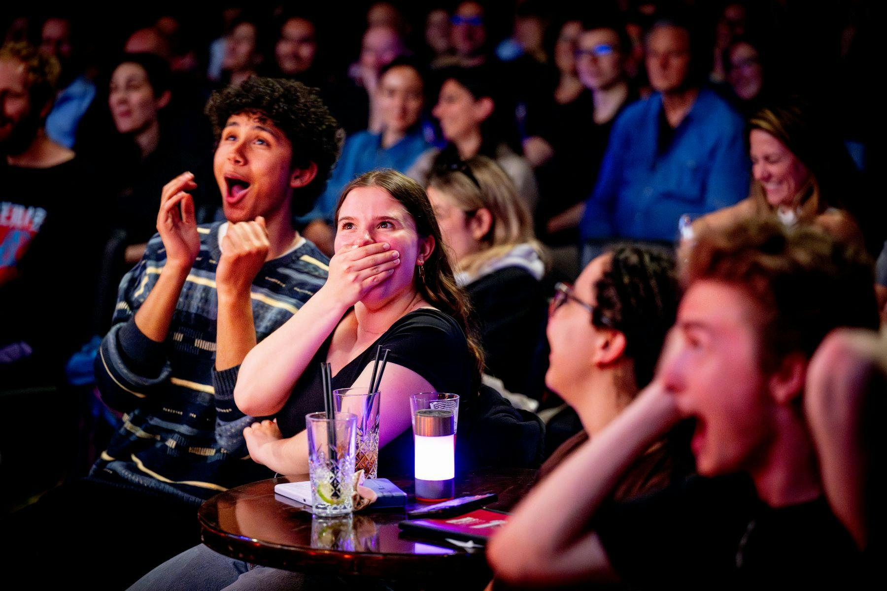 Eine Gruppe von Menschen, die an einem Tisch sitzen und mit Erstaunen und Aufregung reagieren, wobei im Hintergrund ein Publikum zu sehen ist.