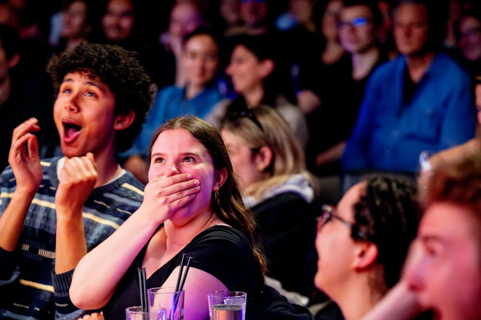 A group of people sitting at a table, reacting with surprise and excitement, with an audience visible in the background.