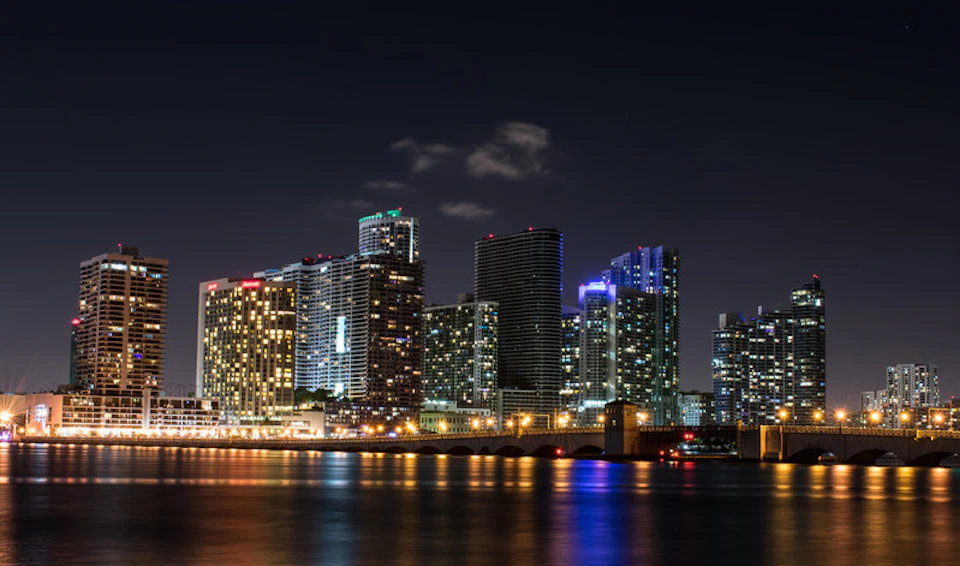 City skyline at night with tall buildings illuminated, reflecting lights on a calm body of water below.