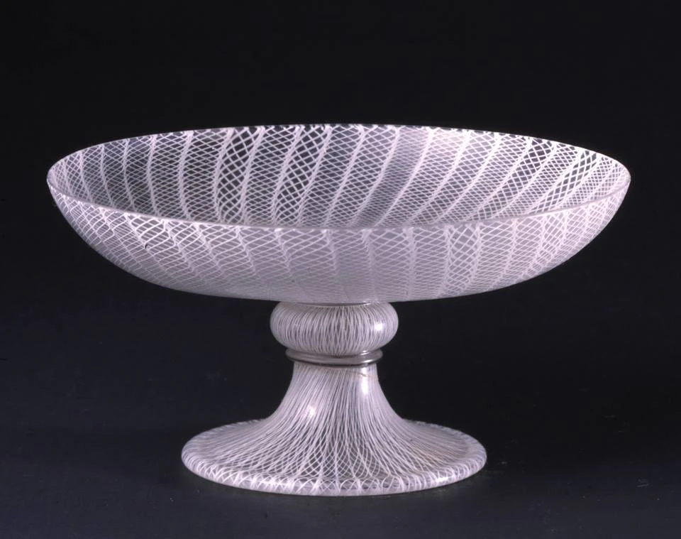 A transparent glass bowl with intricate white lattice patterns sits on a pedestal base against a black background.