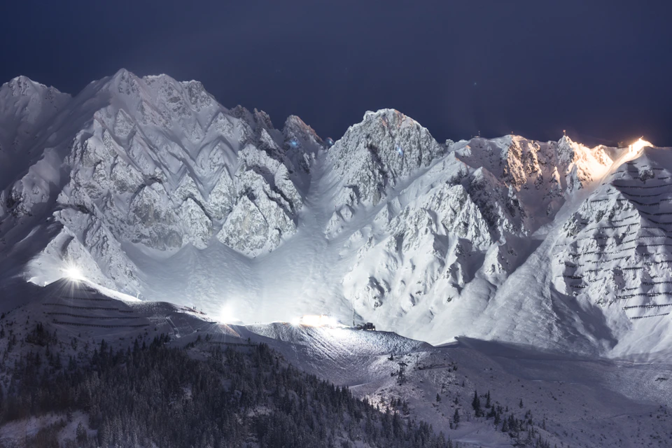 Snow-covered mountain range under a clear night sky, with illuminated ski slopes and pine trees in the foreground.