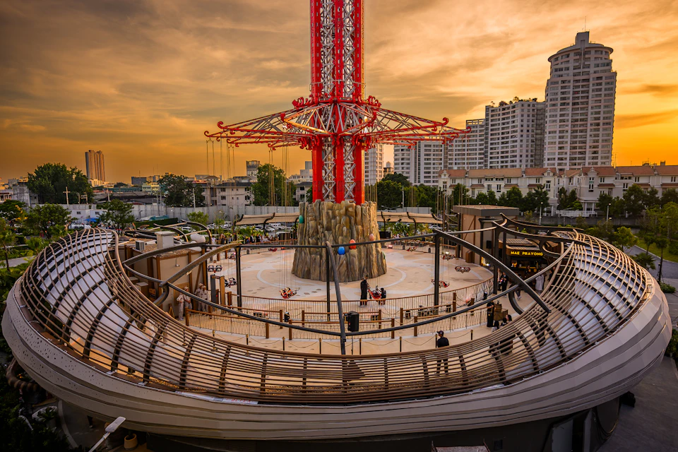 An amusement ride with a tall red structure in an urban area at sunset, surrounded by buildings and a curved railing.