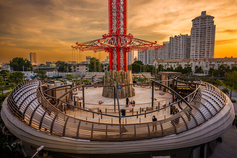 An amusement ride with a tall red structure in an urban area at sunset, surrounded by buildings and a curved railing.