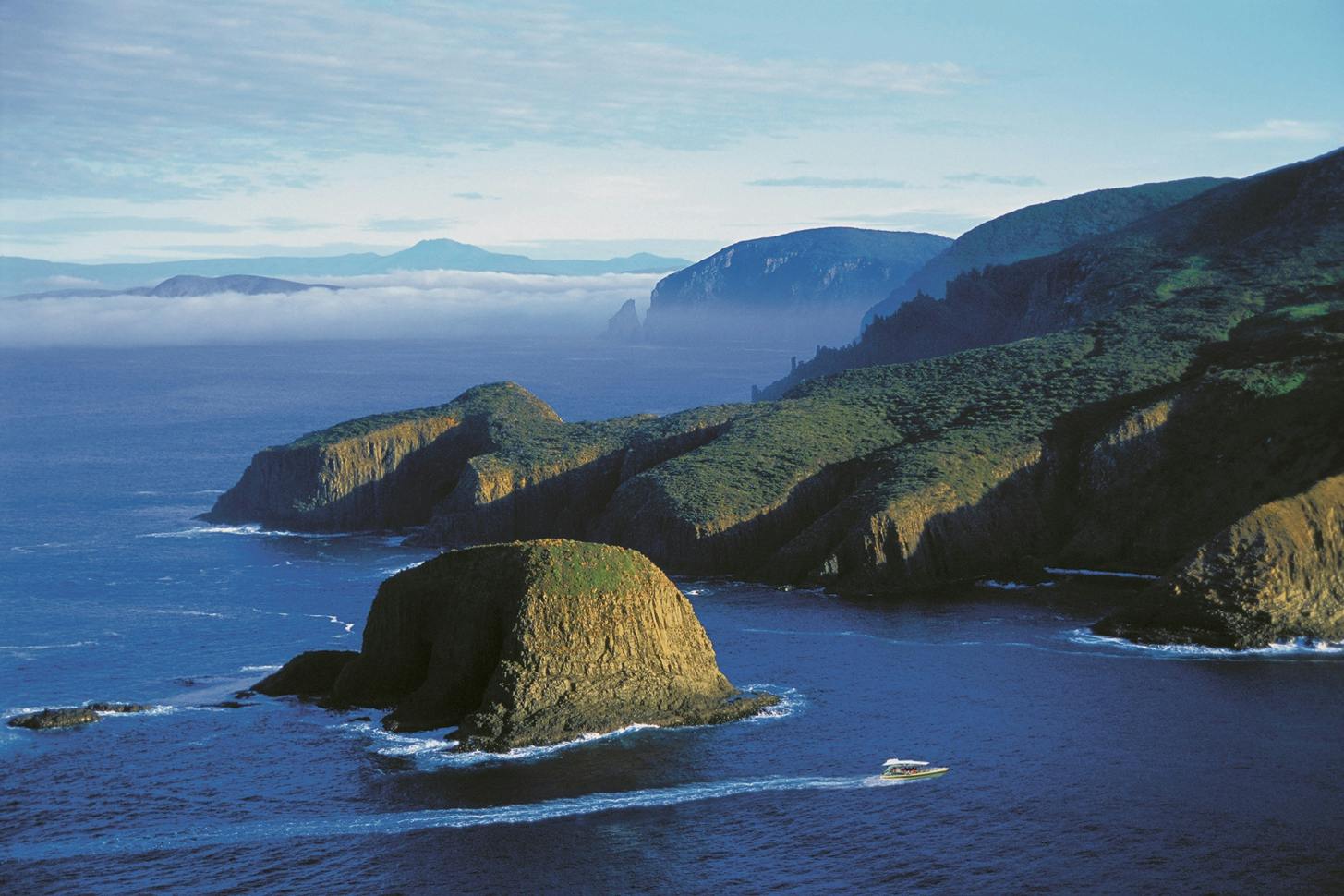 A scenic coastal view with rocky cliffs, green vegetation, and a boat on the water under a blue sky.