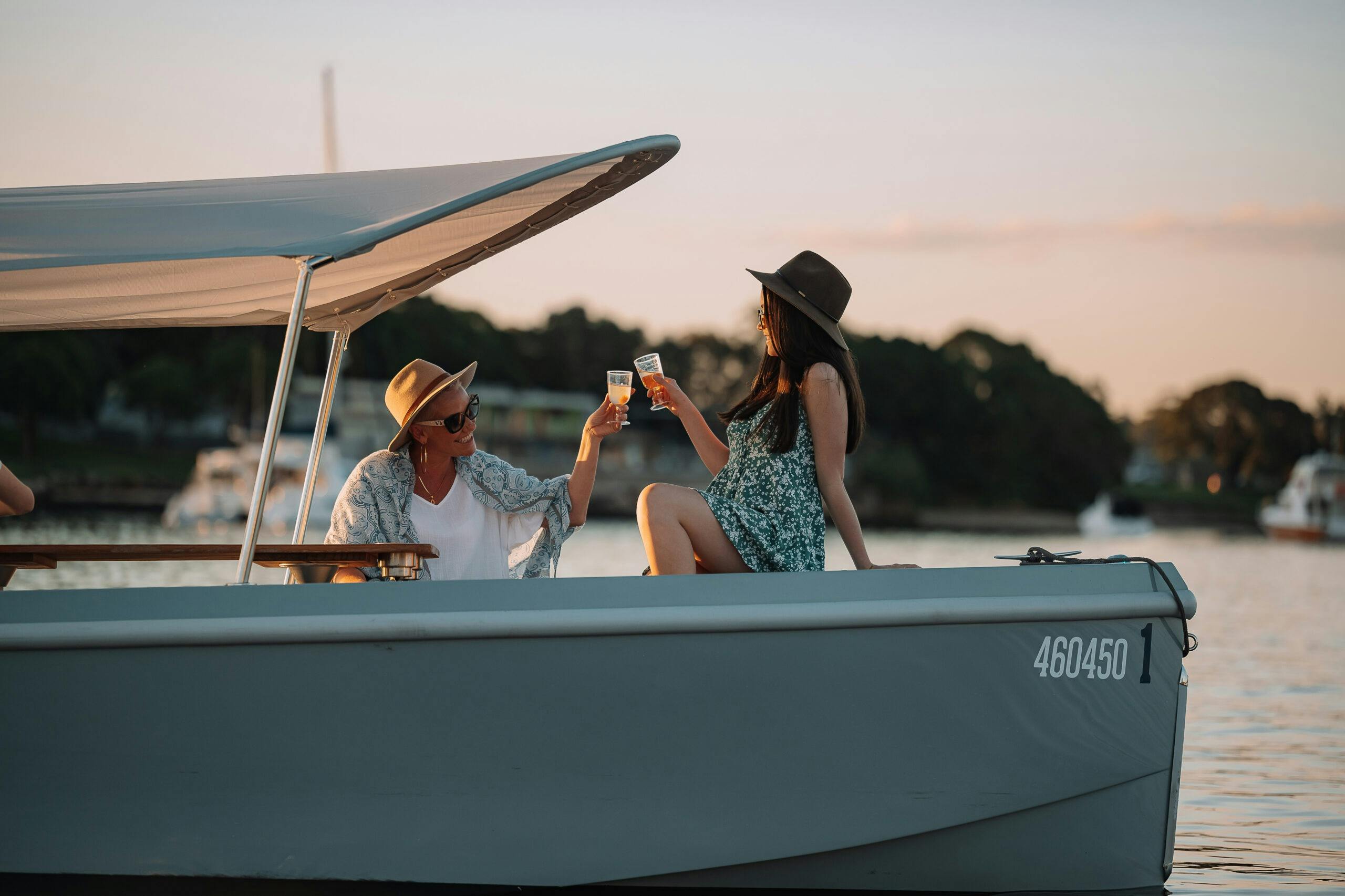 Two people wearing hats toast with drinks on a boat at sunset, with trees and water in the background.