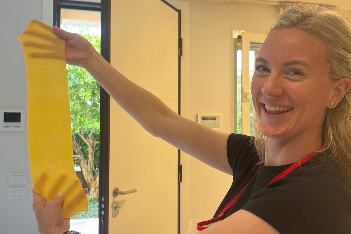 A smiling woman with blonde hair wearing a black shirt and red apron stands indoors, reaching upwards.