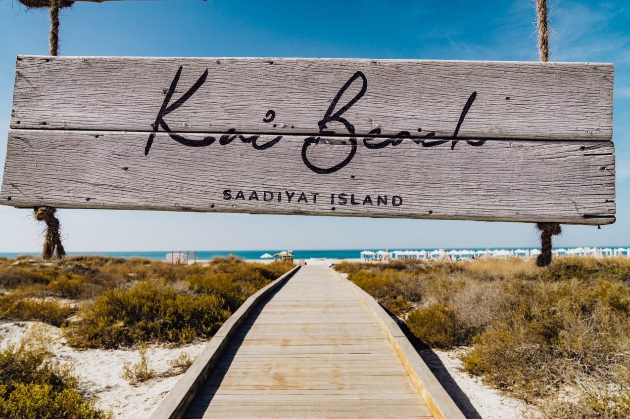 Dřevěná cedule s nápisem „Pláž Kai, ostrov Saadiyat“ nad promenádou vedoucí k oceánu s dunami a vegetací na obou stranách.