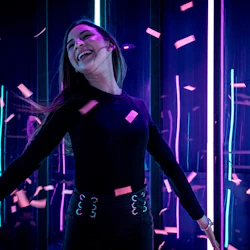 En person dansar glatt bland fallande konfetti, upplyst av livfulla lila och blå neonljus.