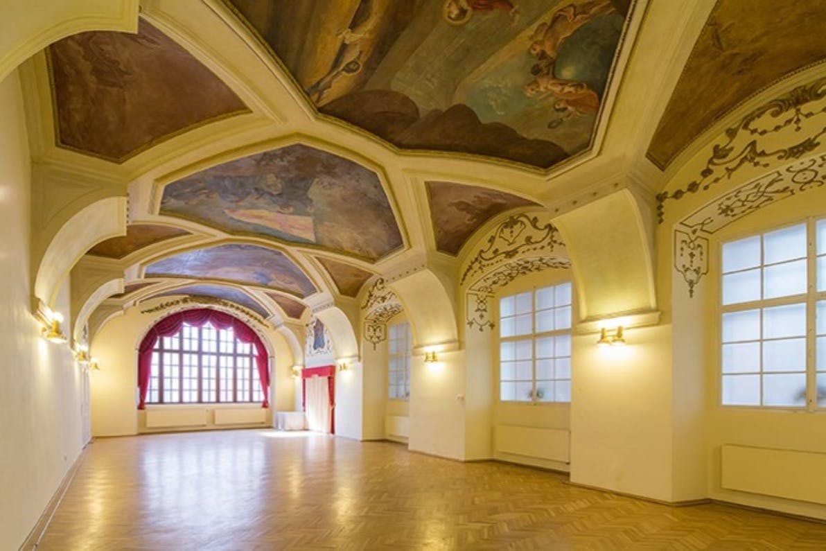 An elegant, spacious hall with ornate painted ceilings, large windows with red curtains, and polished wooden floors.
