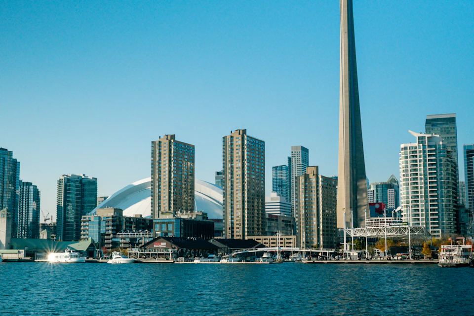 City skyline with tall buildings including the CN Tower, waterfront, and boats on the water under a clear blue sky.