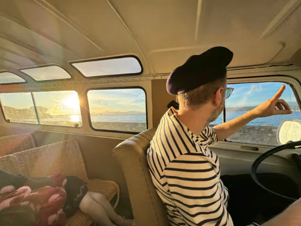 A person in a striped shirt and beret points out of a van window toward the sea, illuminated by the setting sun.