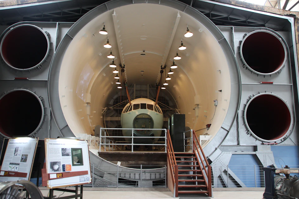 A small aircraft is positioned inside a large, cylindrical testing facility with stairs leading up to it. Two large exhaust vents are on the right.