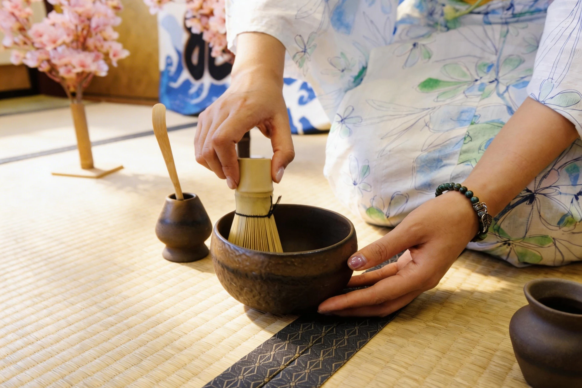 A person preparing matcha tea with a whisk, bowl, and scoop on a traditional mat in a bright room with floral decor.