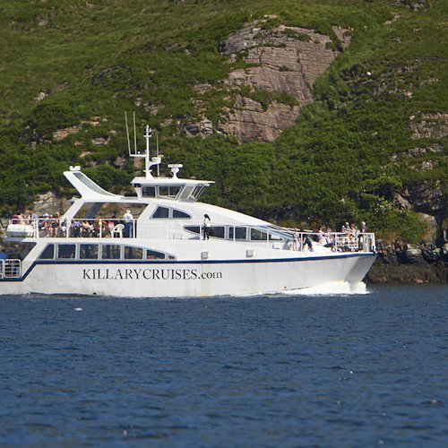 White Killary Cruises boat on water near a green, rocky shoreline, with passengers on the upper and lower decks.