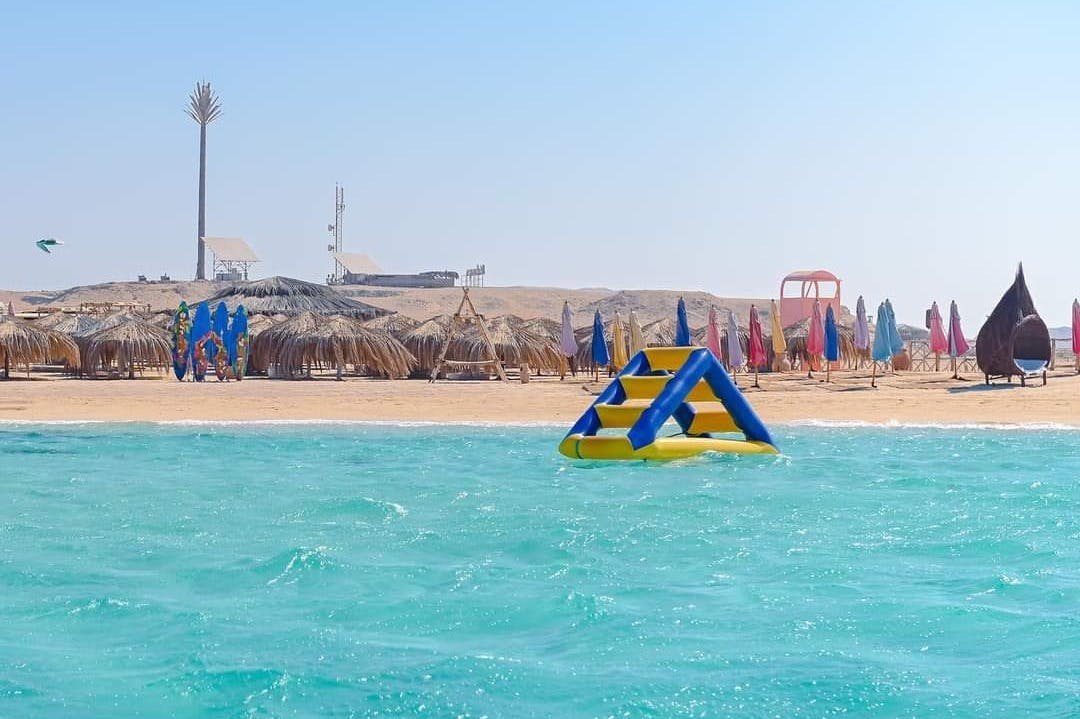 Oppustelig vandstruktur, der flyder i turkis hav med en sandstrand og stråtækte paraplyer i baggrunden.