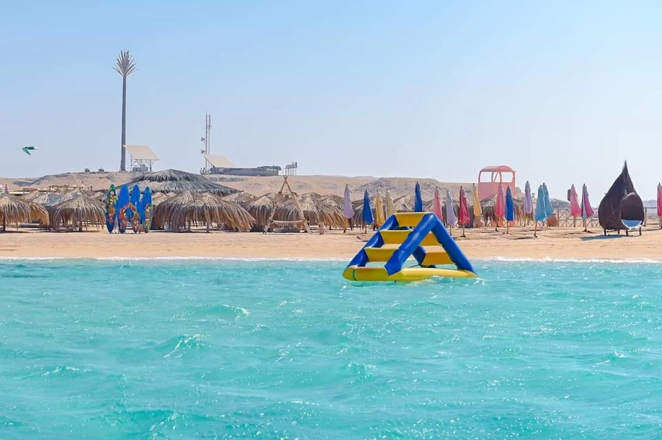 Inflatable water structure floating in turquoise sea with a sandy beach and thatched umbrellas in the background.