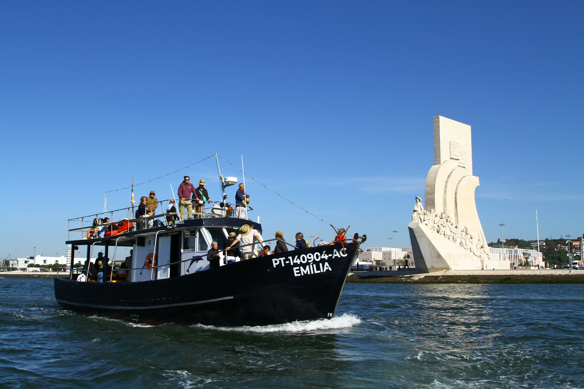 People on a boat named "Emília" sail near a large white monument with sculptures, under a clear blue sky.