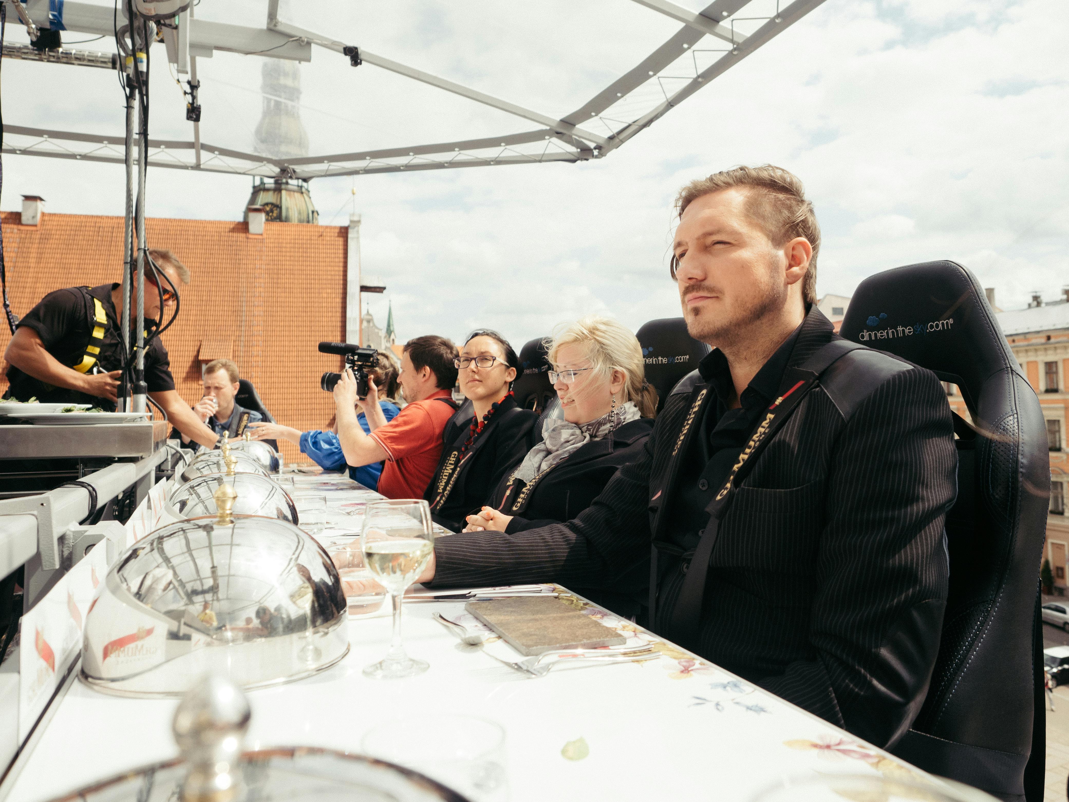 Persones assegudes en una taula de menjador suspesa, algunes amb arnesos de seguretat i una persona filmant amb una càmera.