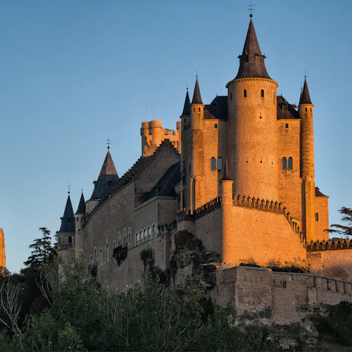 Ett stort medeltida slott med flera torn och en krenelaterad vägg badar i tidigt kvällsljus mot en klar himmel. Träd står i förgrunden.