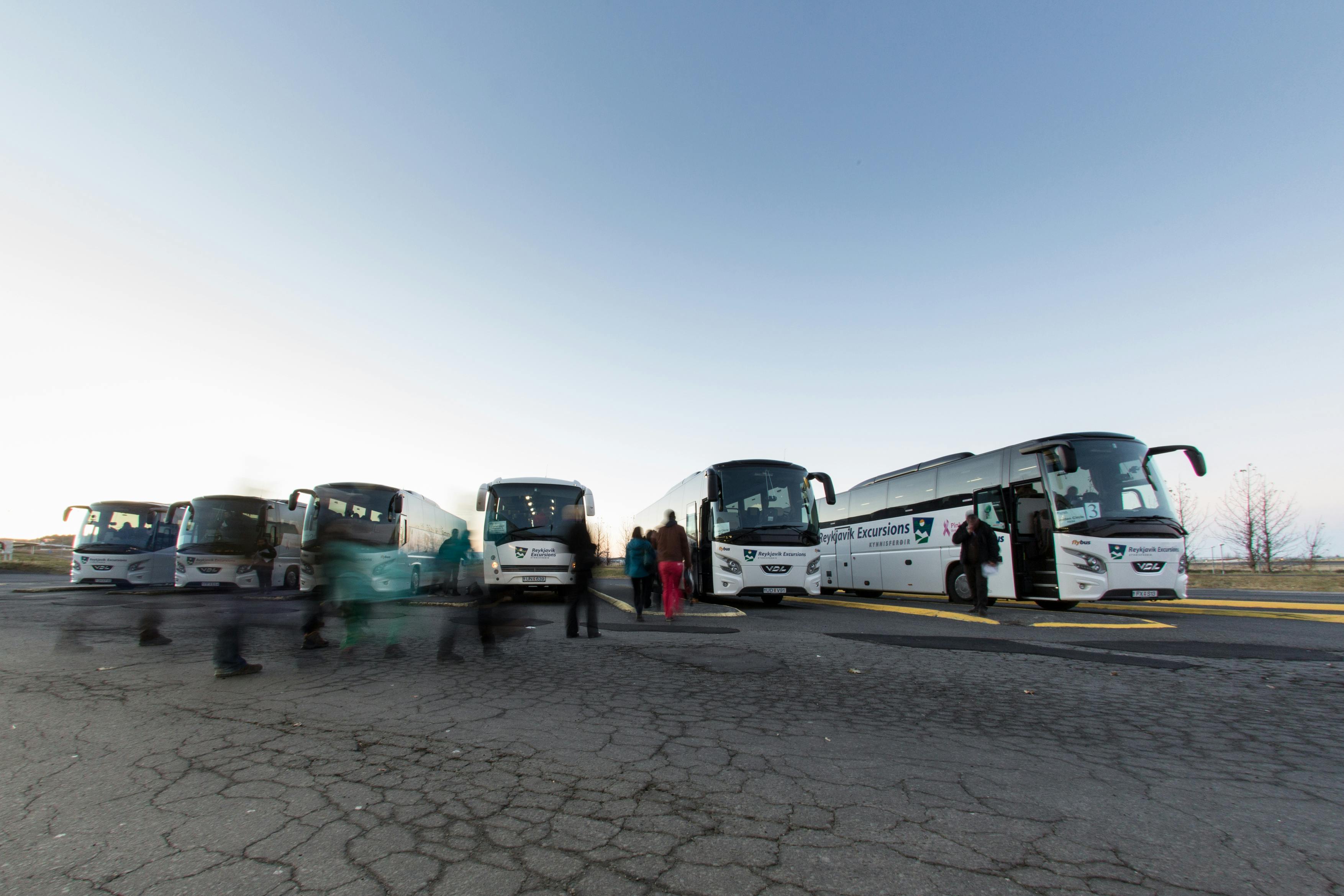 几辆白色旅游巴士停在破碎的人行道上，人们在晴朗的浅蓝色天空下走来走去。