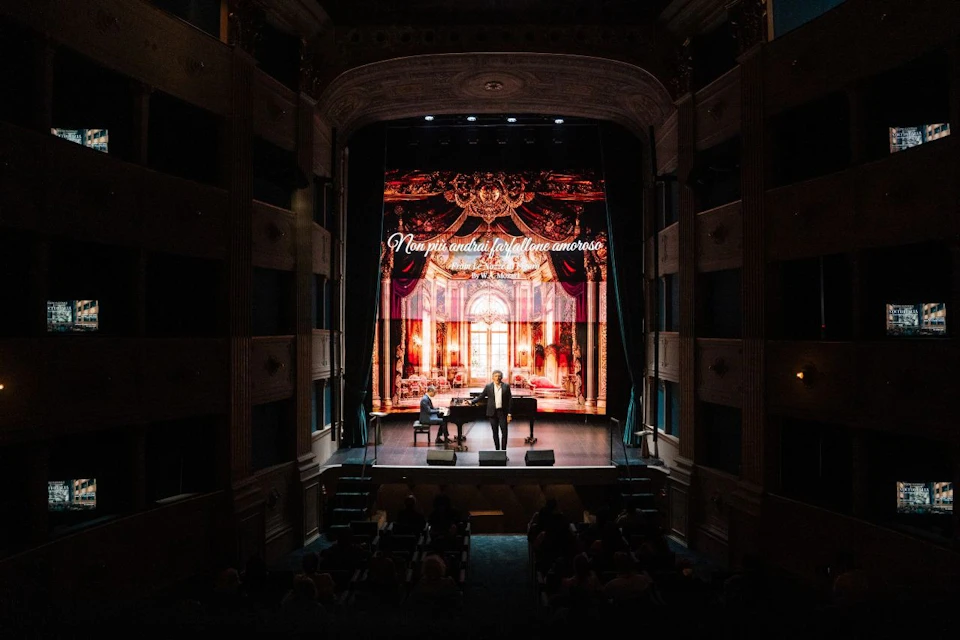 A stage with a performer and a pianist in front of an ornate background image and text, inside a theater with an audience.