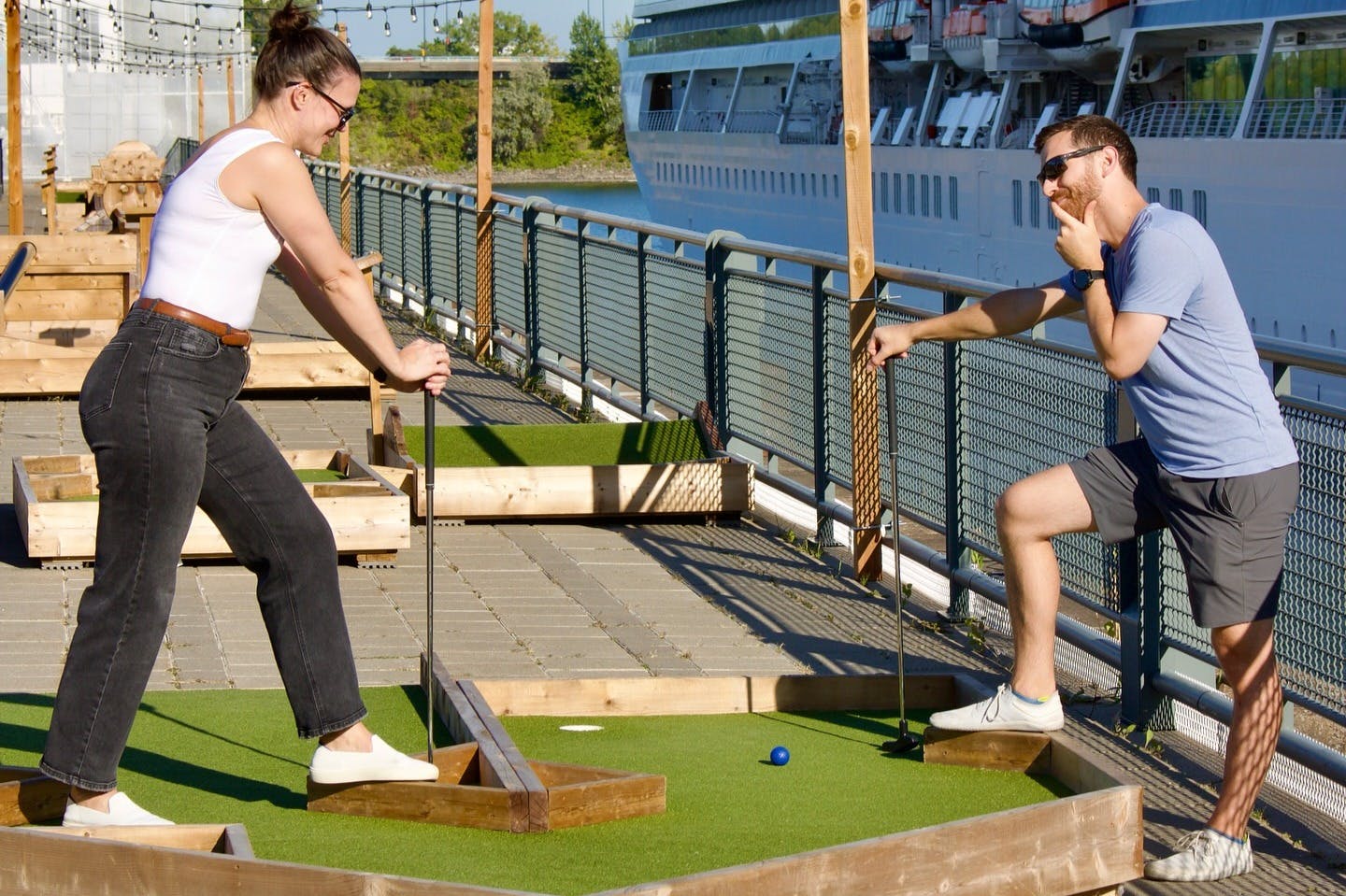 Uma pessoa a jogar mini-golfe num campo exterior junto a um rio, com um grande navio atracado ao fundo.