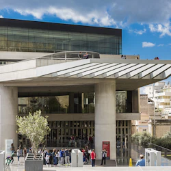 Människor samlas utanför en modern glas- och betongbyggnad med ett stort överhäng, mot en kuliss av urbana strukturer och en delvis molnig himmel.