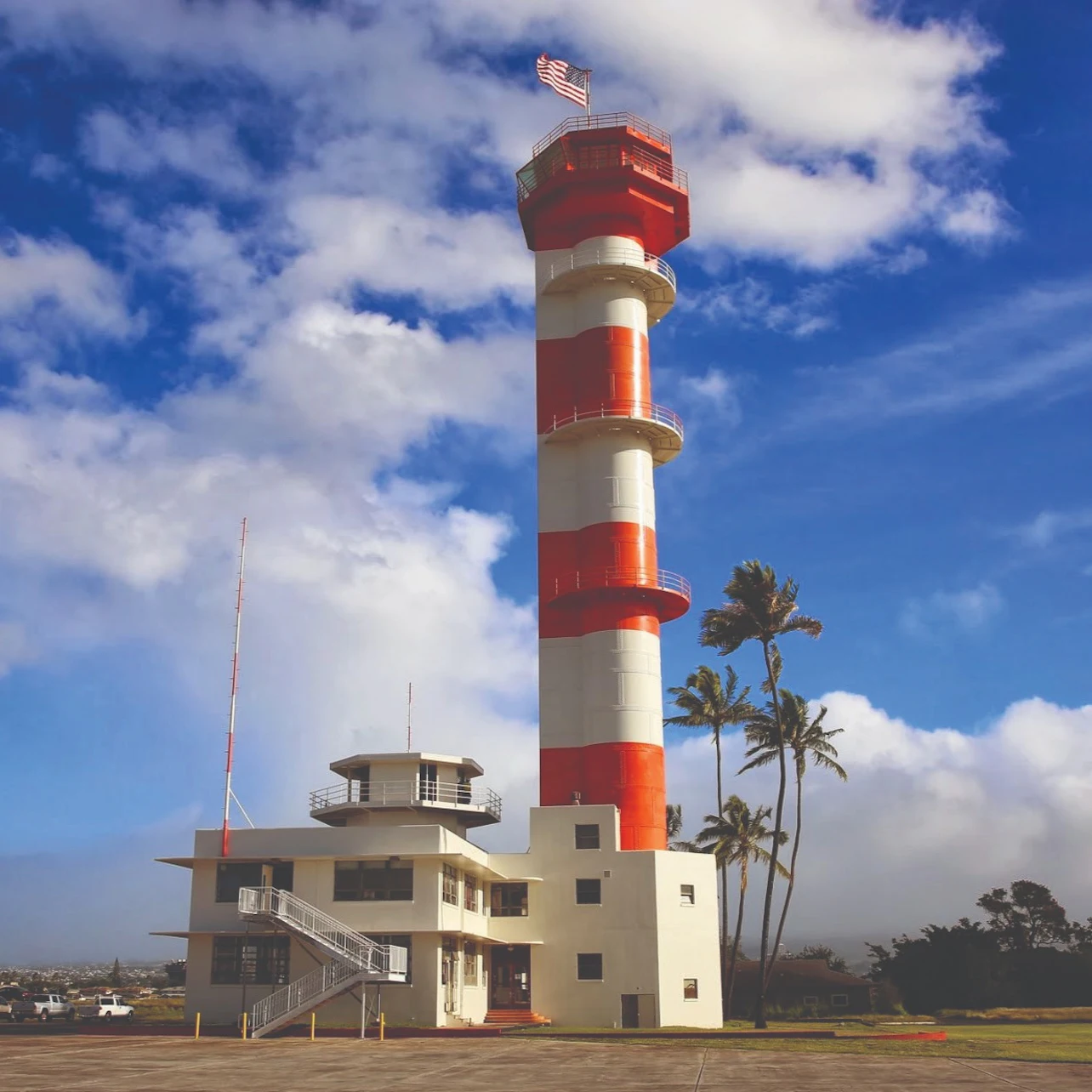 Red-and-white striped control tower with an American flag on top, beside palm trees, under a partly cloudy sky.