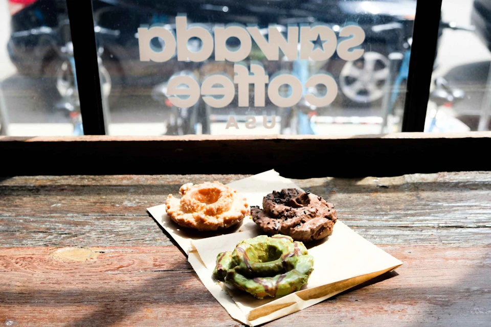 Three assorted crullers on a napkin, placed on a wooden surface in front of a window with "Swaggy Coffee USA" written on it.