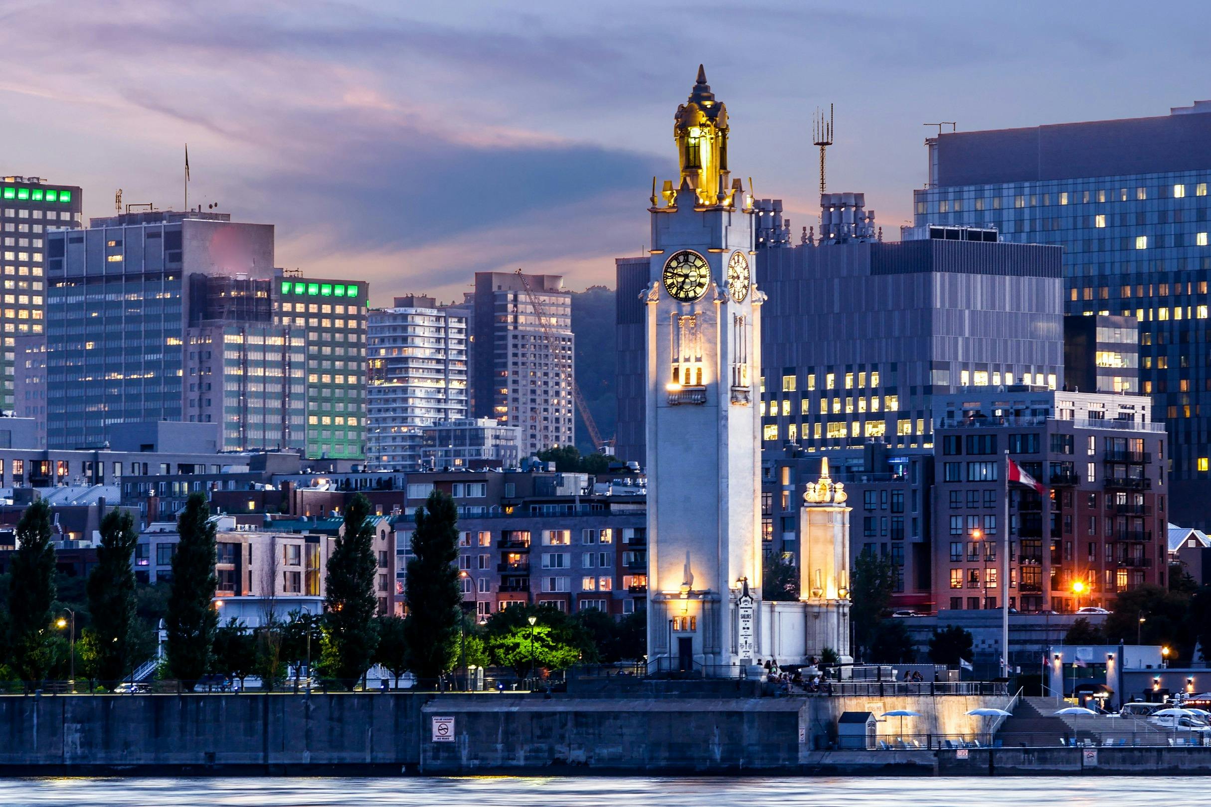 A clock tower illuminated at dusk, surrounded by modern buildings and trees along a waterfront.