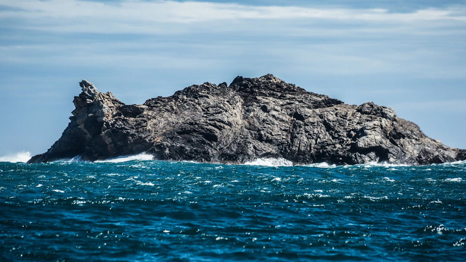 Rough ocean waves crashing against a rocky island under a cloudy sky.