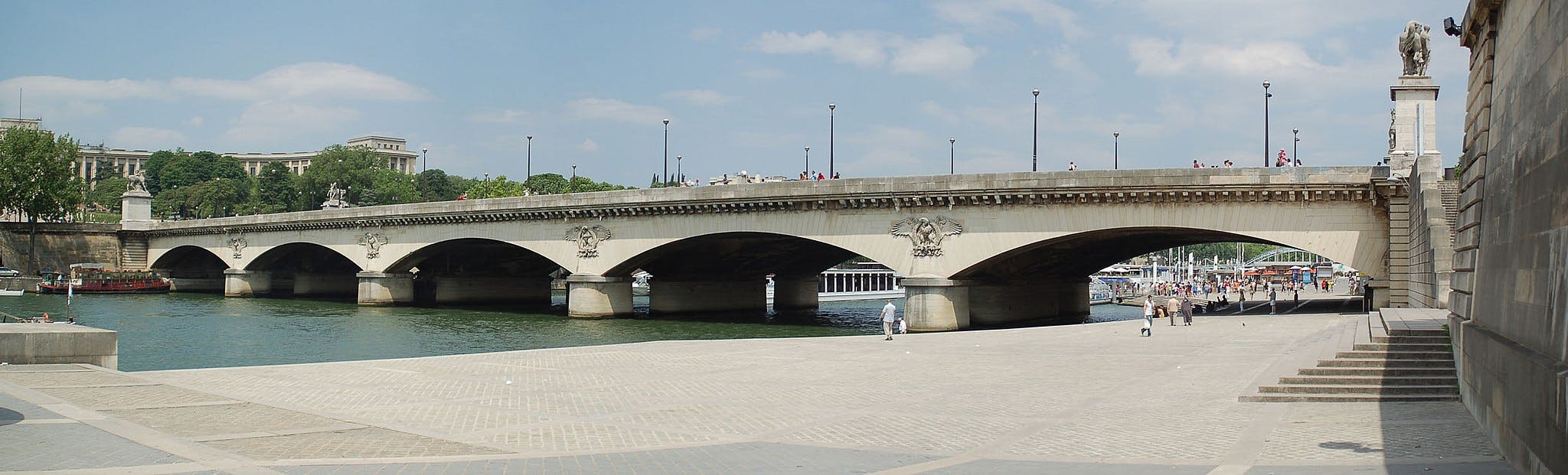 A stone bridge with arches spans a river, with people walking on a pedestrian area beneath. Trees and buildings are visible in the background.