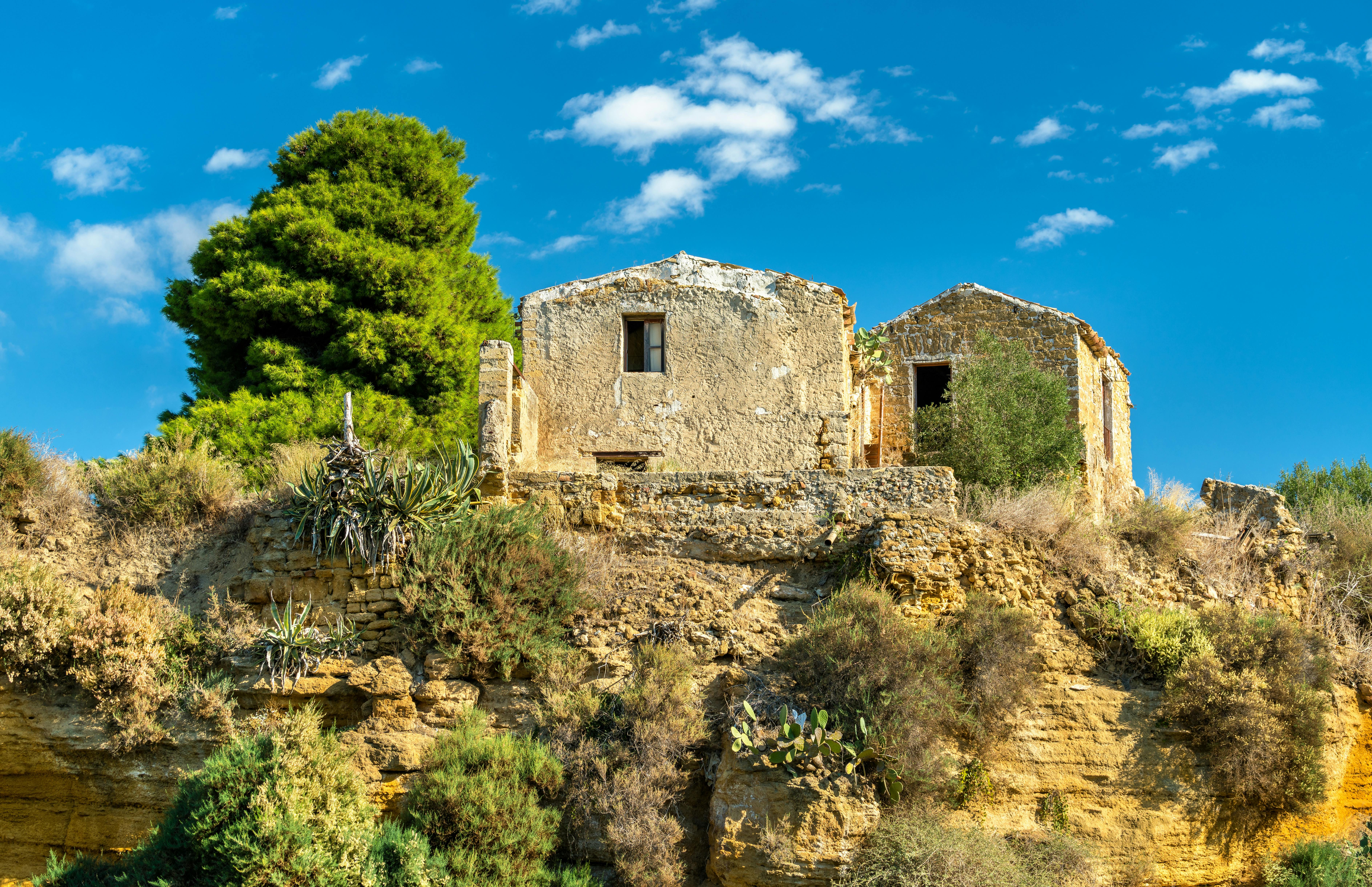 Two old, weathered stone houses sit atop a rocky, vegetation-covered hill under a blue sky with some clouds.